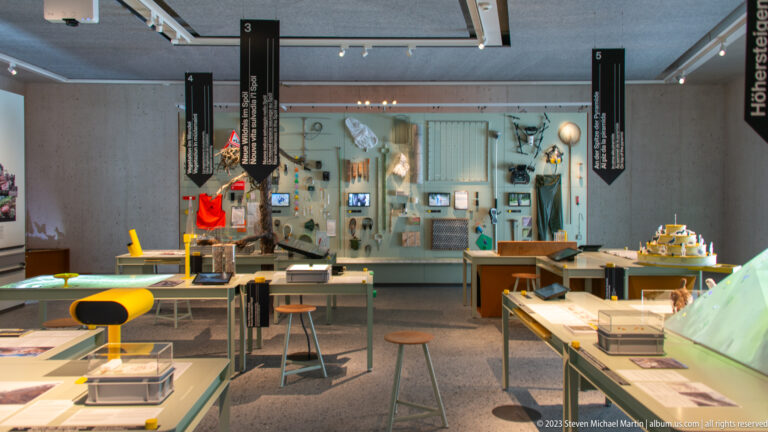 Bright interactive museum gallery with worktables, stools, drawers, suspended panels, models, specimens, and digital displays.