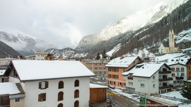 Snowy alpine village with a pale church tower, clustered houses, road, and steep cloud-covered mountains.