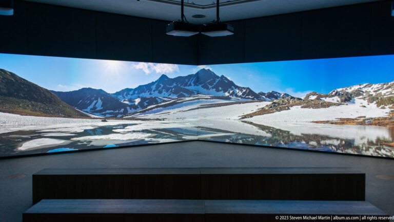 Dark gallery with a wraparound projection of an alpine lake, snowy peaks, and a low viewing bench.