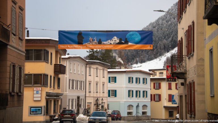 Village street with parked cars and an overhead banner advertising a new exhibition at the national-park center.