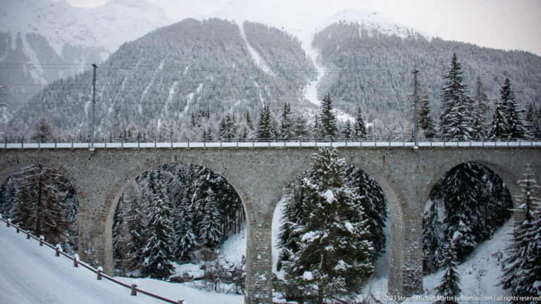 Large multi-arched stone railway viaduct crossing a snow-filled conifer ravine.