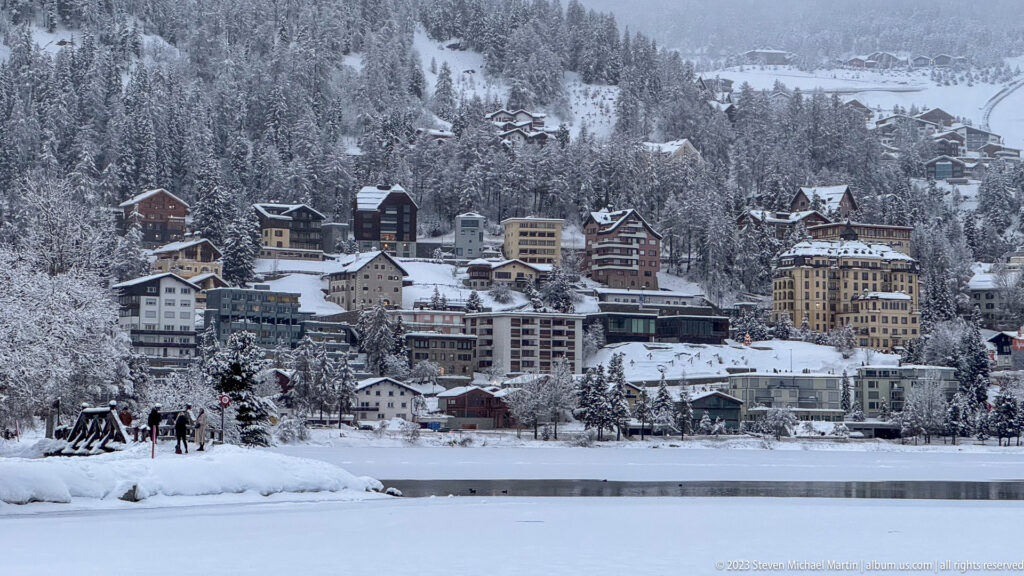 Hotels, apartments, and chalets crowd a snowy St. Moritz hillside above the frozen lake shoreline.