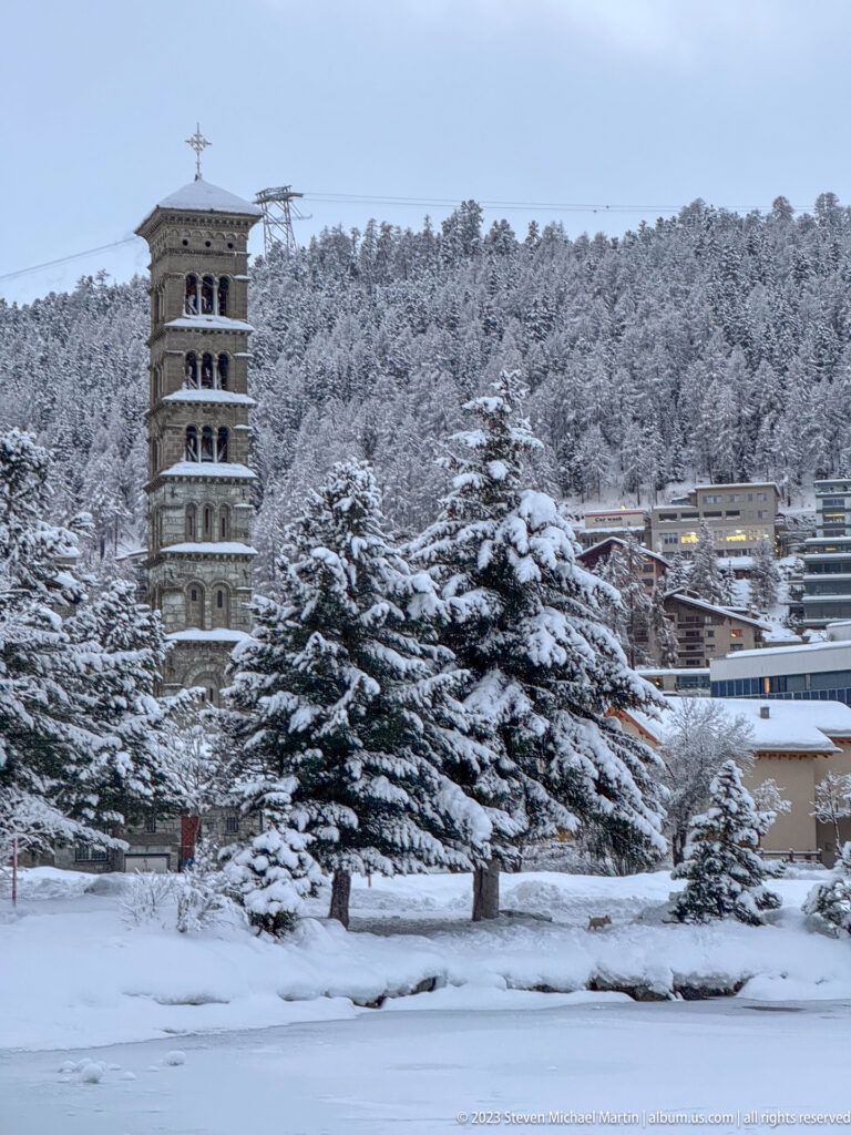 Stone Leaning Tower of St. Moritz rises behind snow-covered evergreen trees, with stacked arched openings visible near the top.