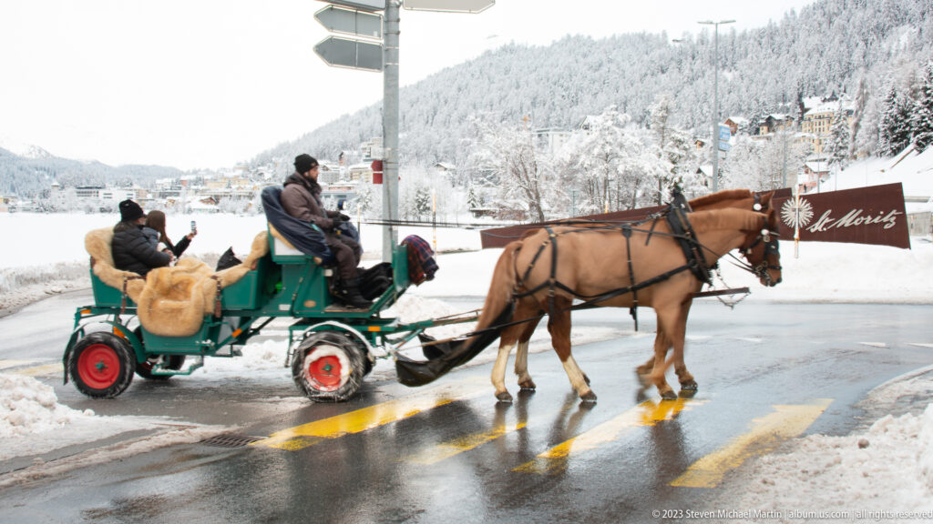 Chestnut horse pulls a green carriage with bundled passengers across a wet, snow-lined street in St. Moritz.