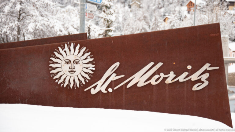 White sun emblem and cursive St. Moritz lettering on a rust-colored metal panel with snow along the bottom edge.
