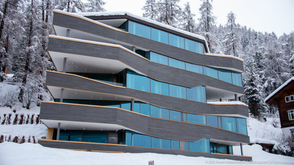 Modern terraced building with dark cladding and blue-tinted windows stands beside snow-covered conifer forest.