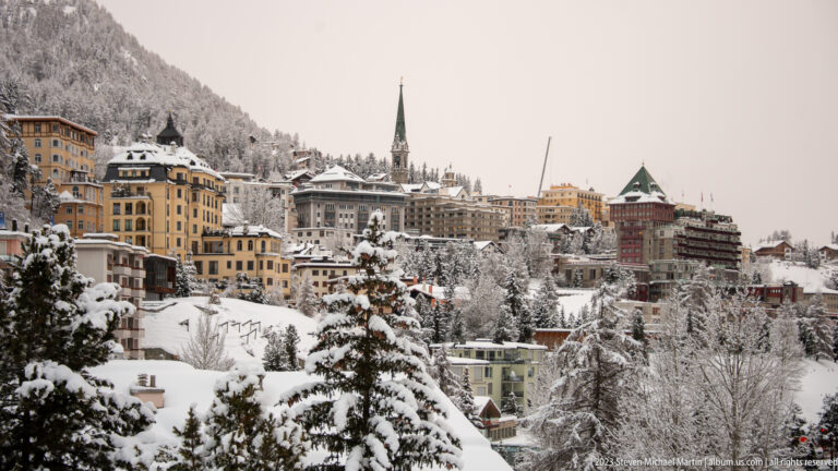 Hotels and houses crowd a snowy St. Moritz hillside around a tall, narrow church spire.