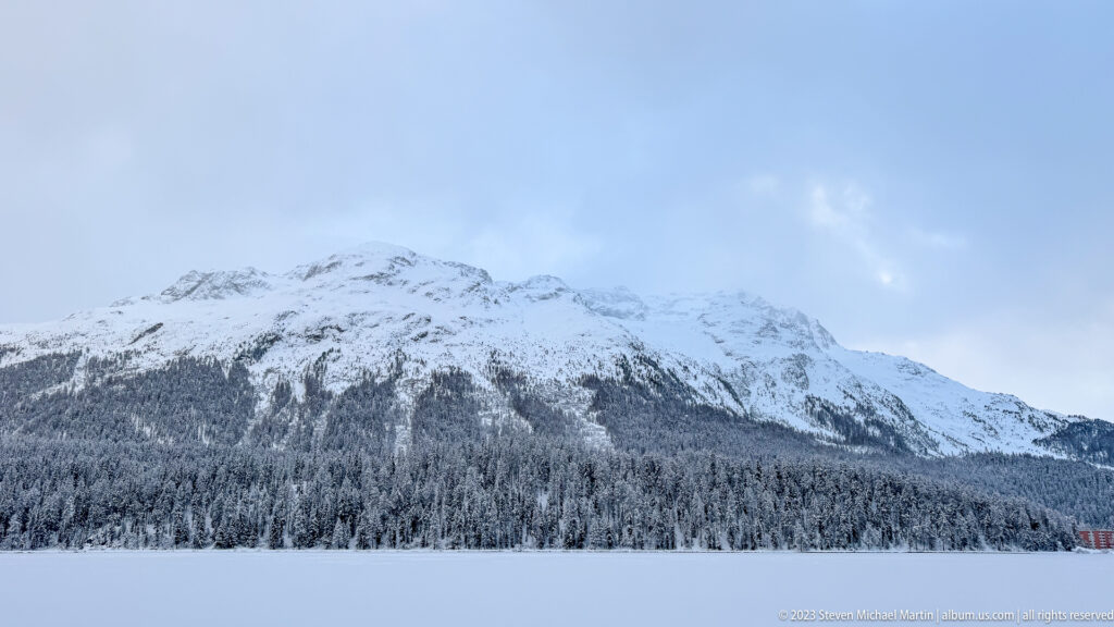 Snow-striped mountain rises above dense conifer forest and the flat white surface of frozen Lake St. Moritz.