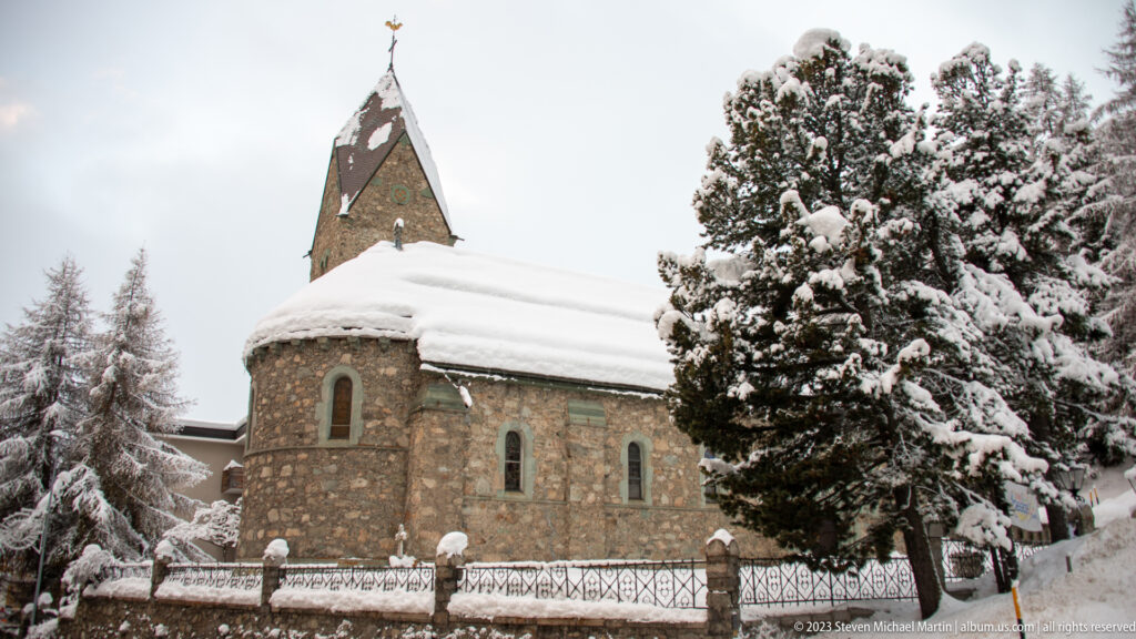 Rounded stone church apse and steep tower rise beside snow-covered conifer trees and a low churchyard wall.