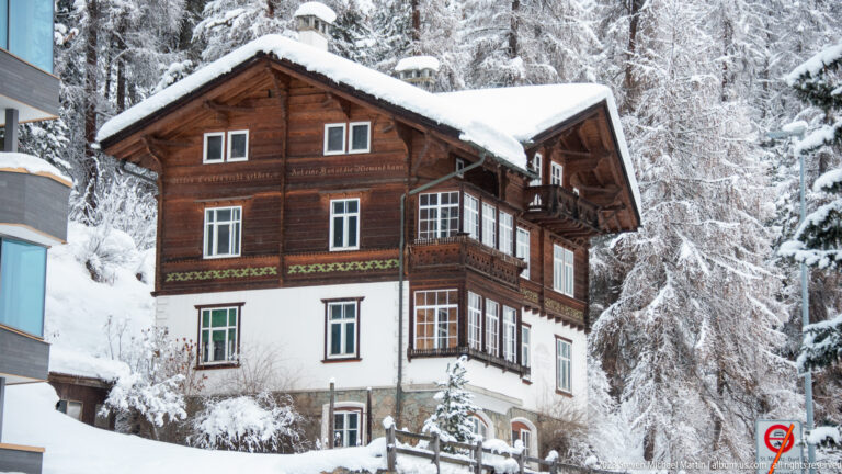 Traditional Alpine house with white stucco lower walls, dark timber upper stories, balconies, and a snow-covered roof.