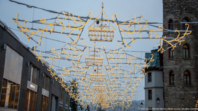 Warm-white Christmas lights form a branching overhead canopy with repeated geometric motifs above a St. Moritz street.