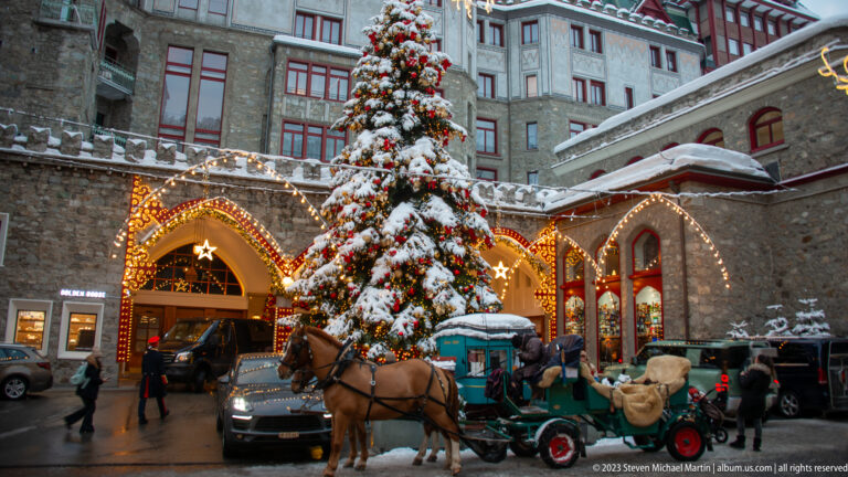 Horse-drawn carriage stands beside a large decorated Christmas tree and illuminated stone-arched hotel entrance in the snow.