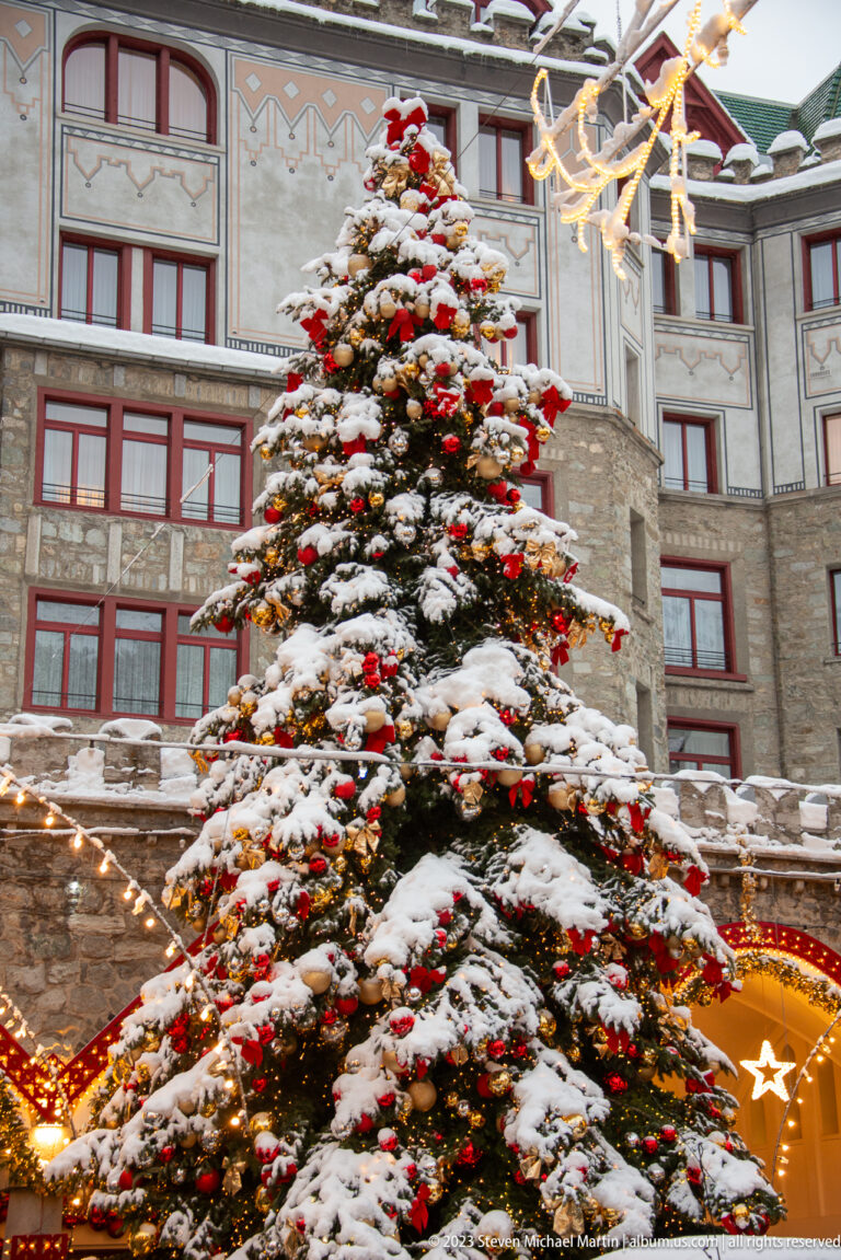 Large Christmas tree covered in snow, red and gold ornaments, and lights stands before a multistory St. Moritz building.