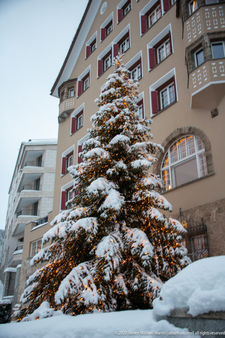 Lit, snow-covered Christmas tree rises in front of a hotel façade with red shutters, balconies, and an arched opening.