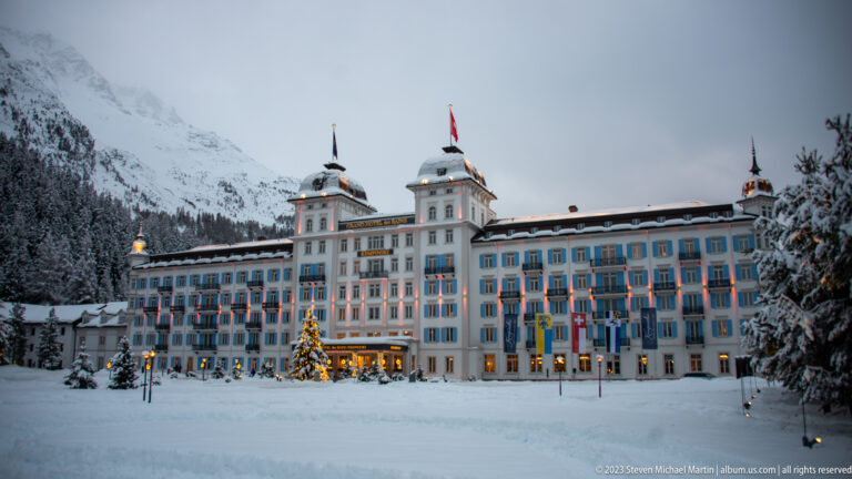 Grand Hotel des Bains Kempinski glows at dusk behind deep snow, with blue shutters and a lit Christmas tree.