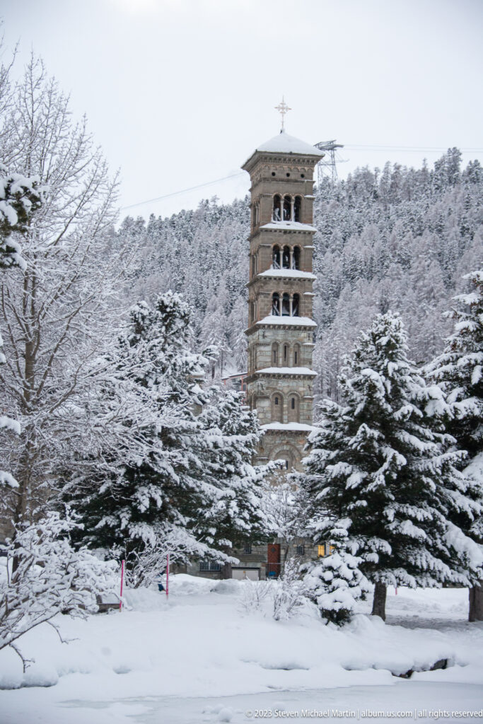 Leaning Tower of St. Moritz rises behind snow-covered trees, with its stacked open arcades clearly visible.
