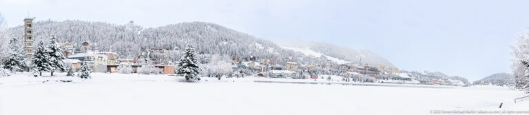 Snow-covered St. Moritz buildings rise on a wooded hillside beyond the frozen lake under low winter clouds.