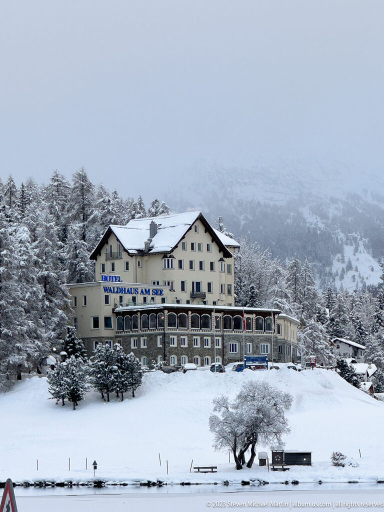 Hotel Waldhaus am See stands above a snowy lakeshore with a wooded mountain slope behind it.