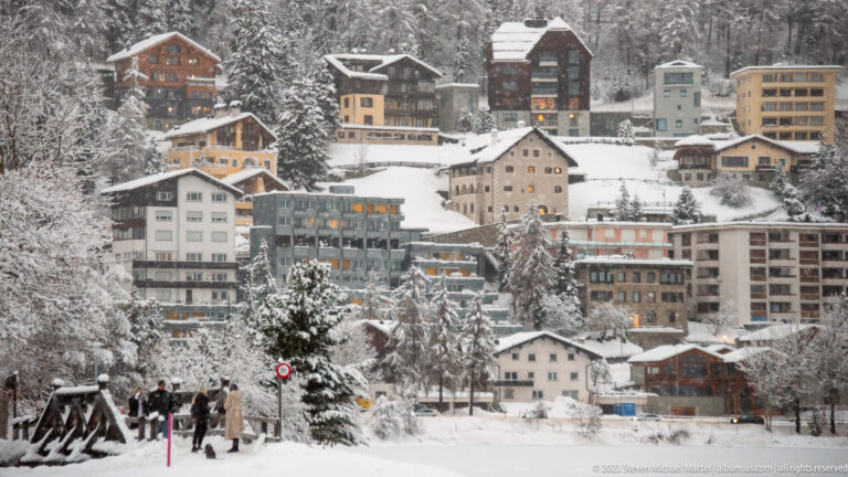 Snow-covered chalets, apartments, and hotel buildings cluster tightly on a steep wooded St. Moritz hillside.
