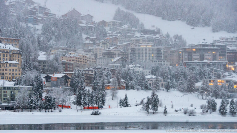 Snow-covered buildings and chalets climb a wooded St. Moritz hillside above the lake, with scattered windows glowing in winter light.
