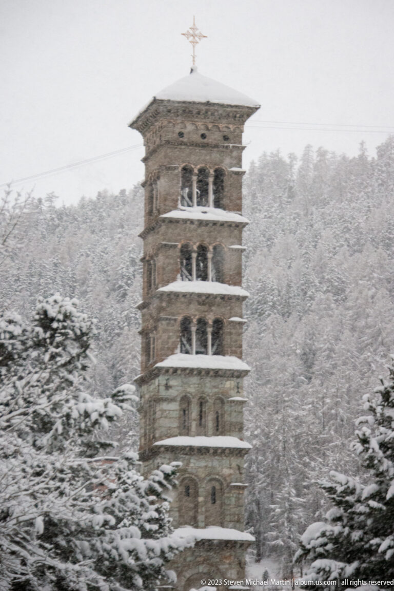 Snow-covered stone bell tower with stacked arched openings and a weather vane, backed by a dense forest on a snowy hillside.