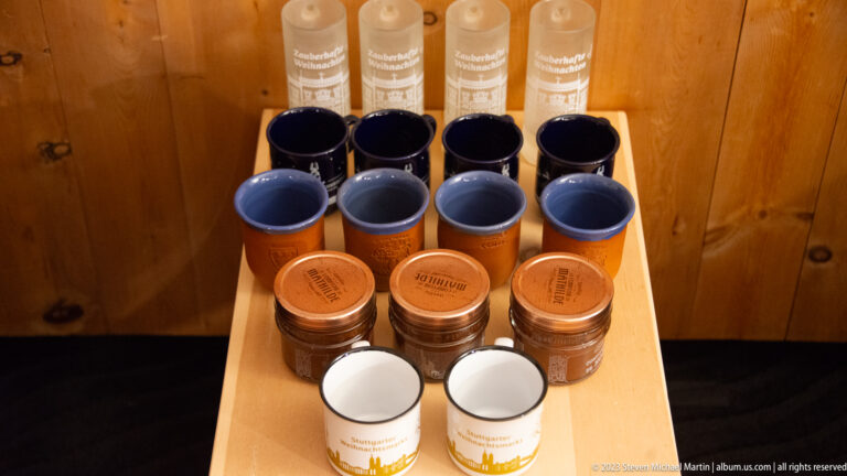 Rows of dark and light ceramic mugs, lidded containers, and tall cylindrical packages arranged on a small wooden display.