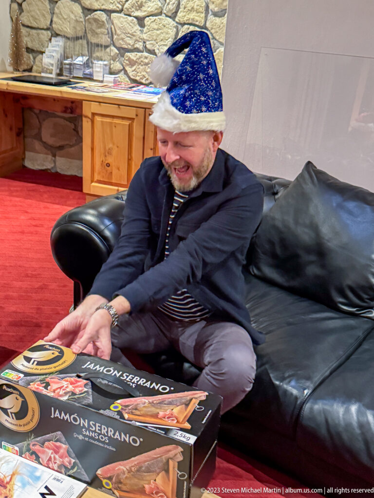 Smiling man in a blue Santa hat seated on a black leather sofa beside a boxed jamón serrano set.