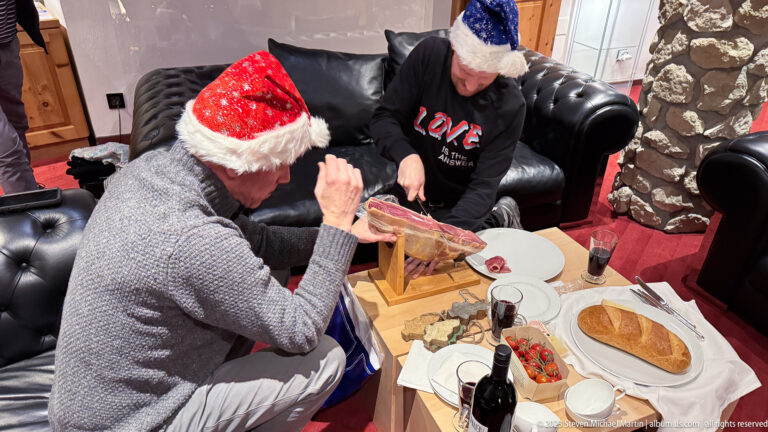 Two men in Santa hats sit around a low table with mounted cured ham, bread, cheese, tomatoes, wine, and plates.