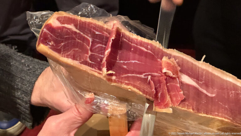 Close-up of a cured ham being sliced with a long knife, showing red meat, white fat, and the pale rind.