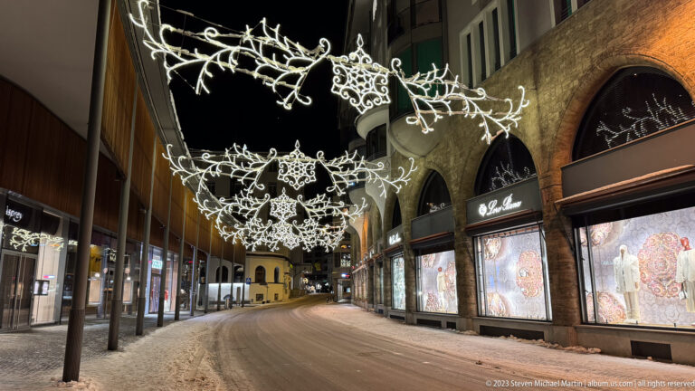 Snowy St. Moritz shopping street at night beneath ornate white Christmas lights, with illuminated storefronts and arched façades.