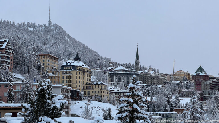 Snowy hotels and houses climb the St. Moritz hillside around a tall church spire and dark conifer trees.