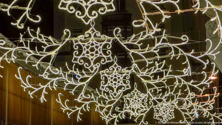 Close-up of ornate white Christmas lights forming a central snowflake with curling branches and scrollwork against a dark façade.