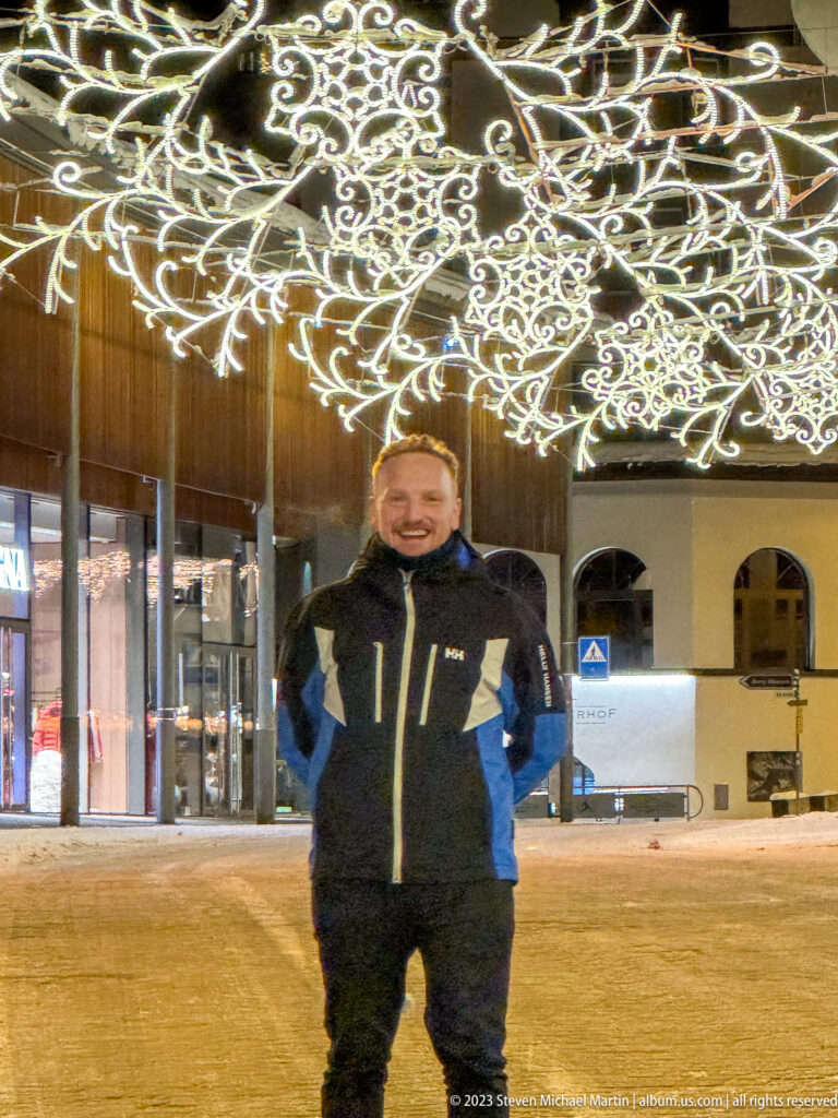 Man in a winter jacket standing on a snowy street beneath a broad canopy of ornate white Christmas lights.