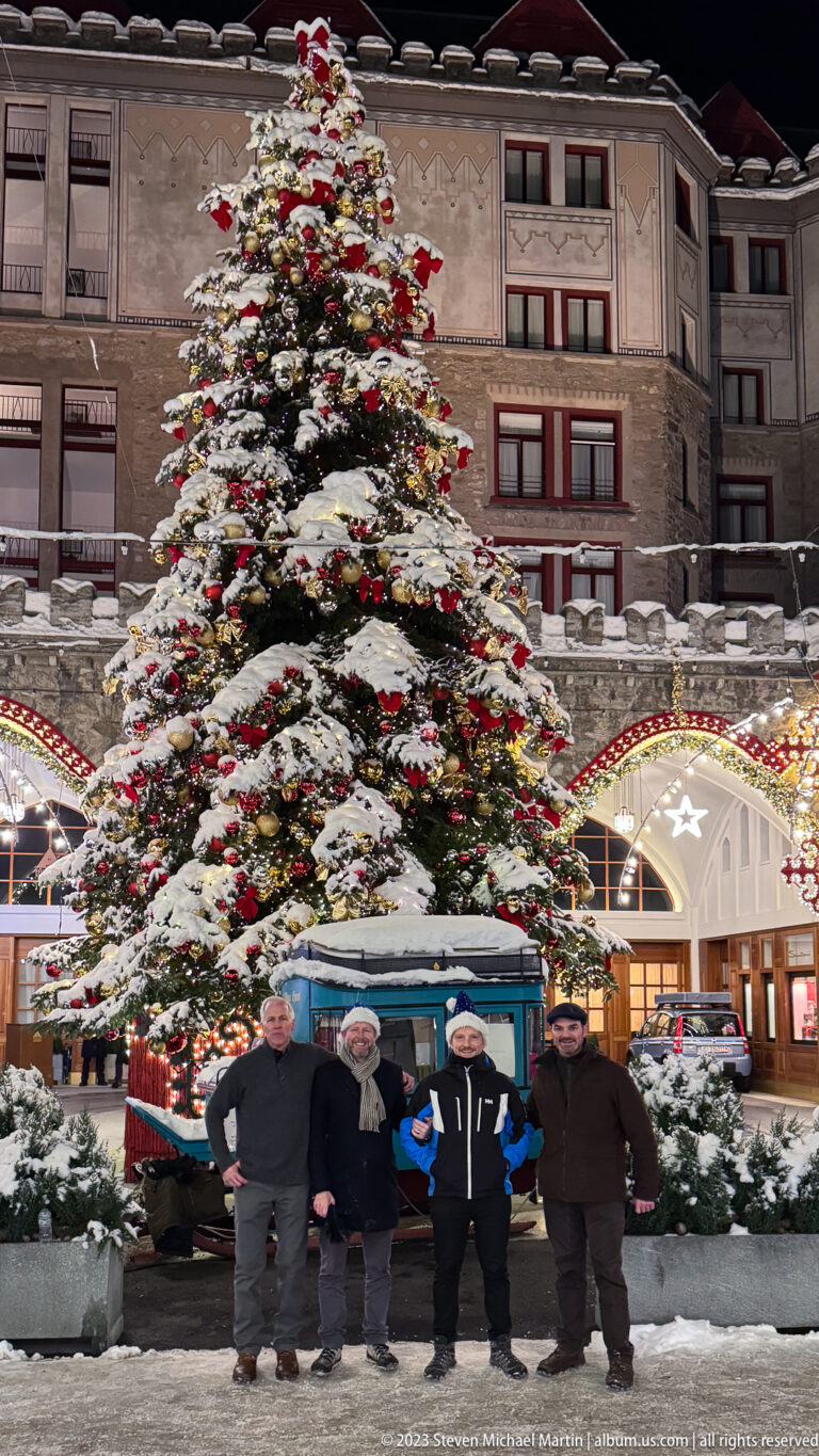 Four men pose in snow before a large decorated Christmas tree with red bows, gold ornaments, and a blue display vehicle behind.