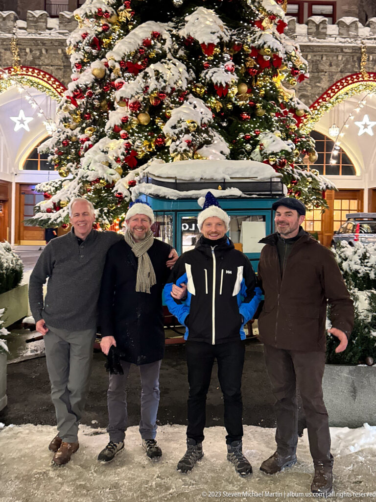 Four men pose beside a blue vehicle marked Palace Hotel, with a snow-covered decorated Christmas tree behind them.