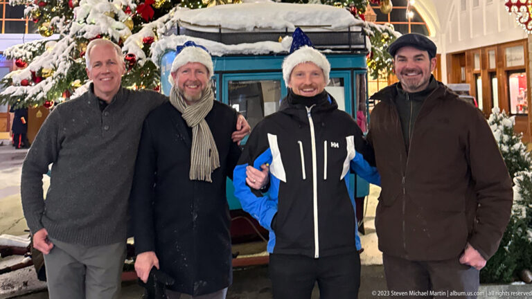 Four men pose closely together, two in festive hats, with a blue Palace Hotel display and Christmas tree behind.