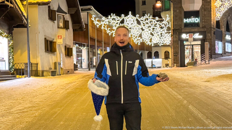 Man on a snowy St. Moritz street holding a blue Santa hat beneath glowing Christmas decorations at night.