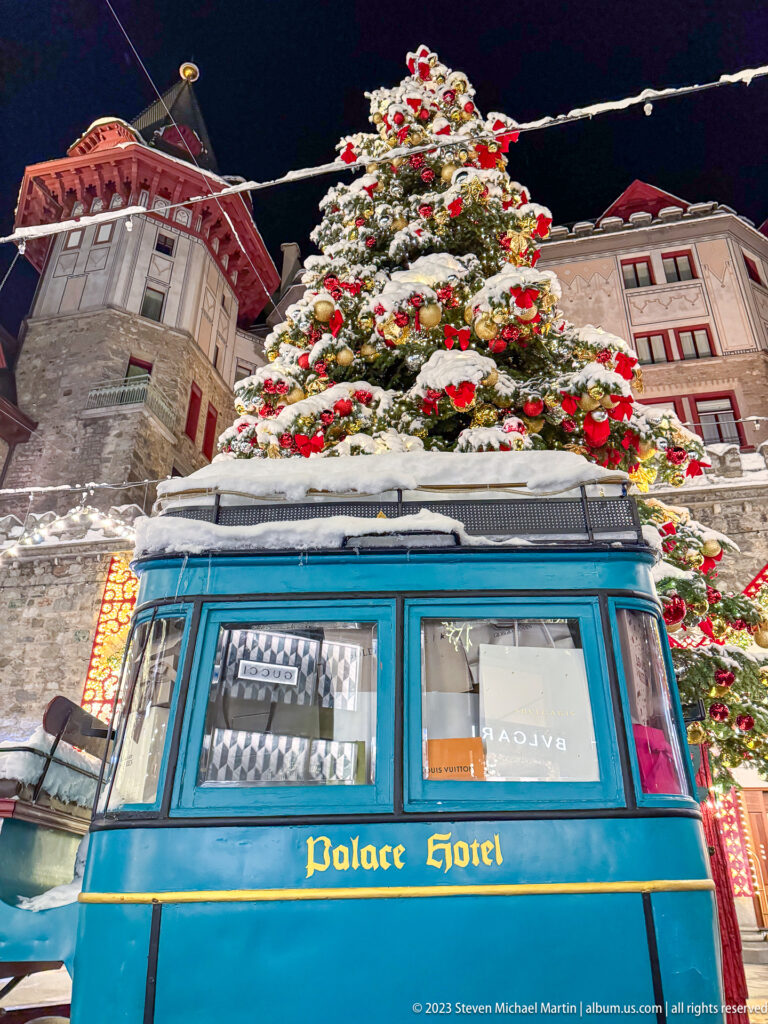 Close low-angle view of a blue Palace Hotel vehicle beneath a snow-covered Christmas tree and historic hotel façade.