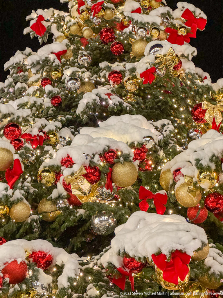 Close-up of snowy Christmas tree branches decorated with red bows, gold baubles, reflective ornaments, and warm white lights.
