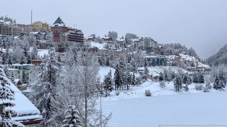 Snow-covered buildings and chalets climb the wooded St. Moritz hillside beyond the frozen lake.