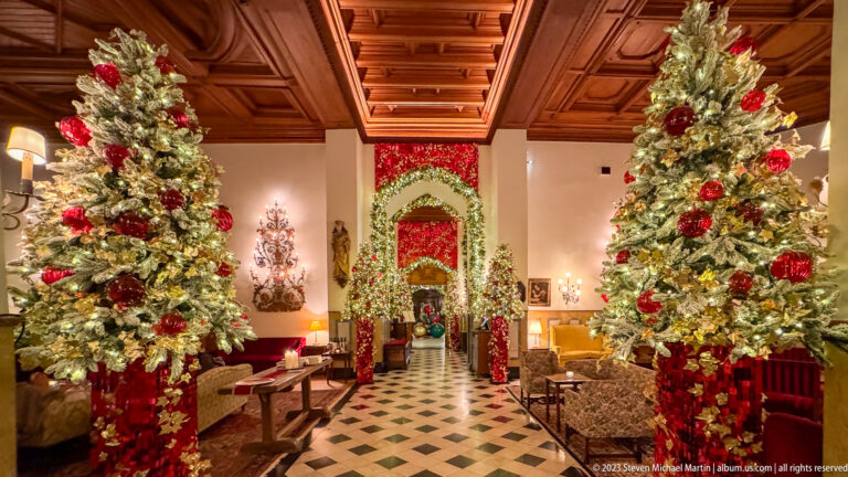 Formal hotel lobby with two decorated Christmas trees, garlanded archways, patterned tile floor, seating, and a coffered wood ceiling.