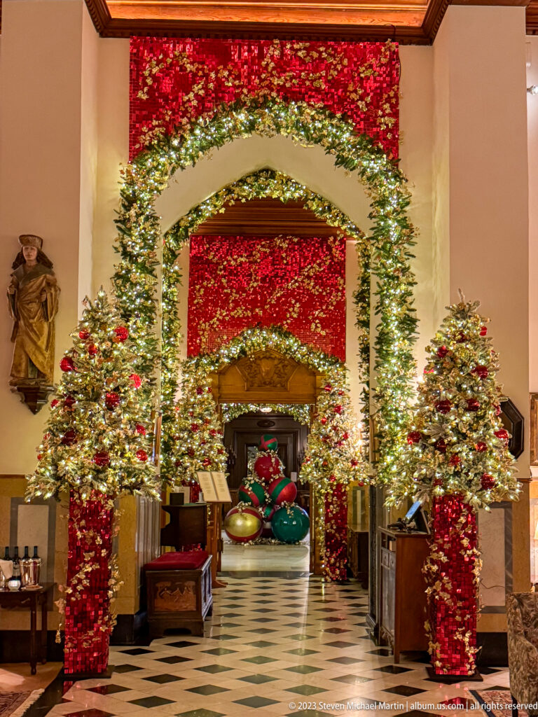 Garland-covered arches frame a hotel passage with two small Christmas trees and oversized ornaments visible deeper inside.