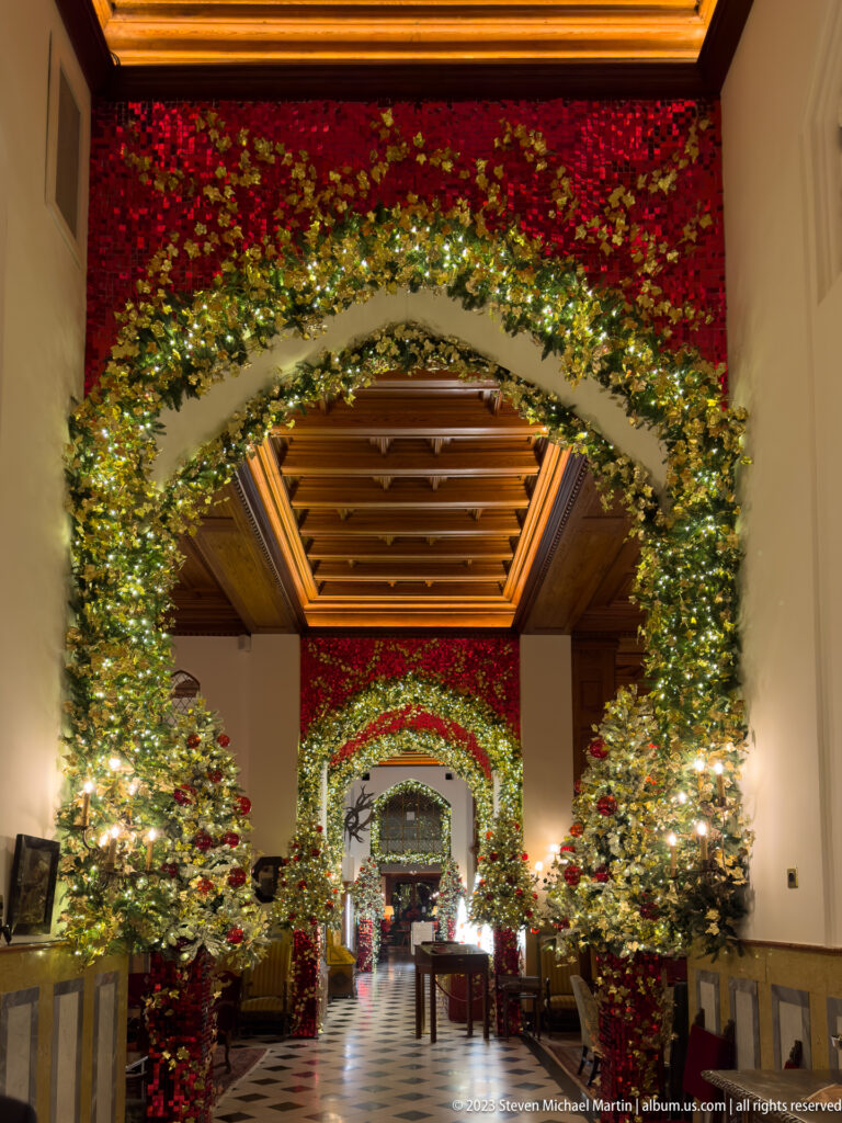 View through a hotel lobby framed by glowing garland arches, paired Christmas trees, and a dark coffered wood ceiling.