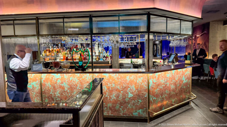 Modern bar with a glowing faceted counter, shelves of liquor bottles, hanging wine glasses, bartenders, and patrons.