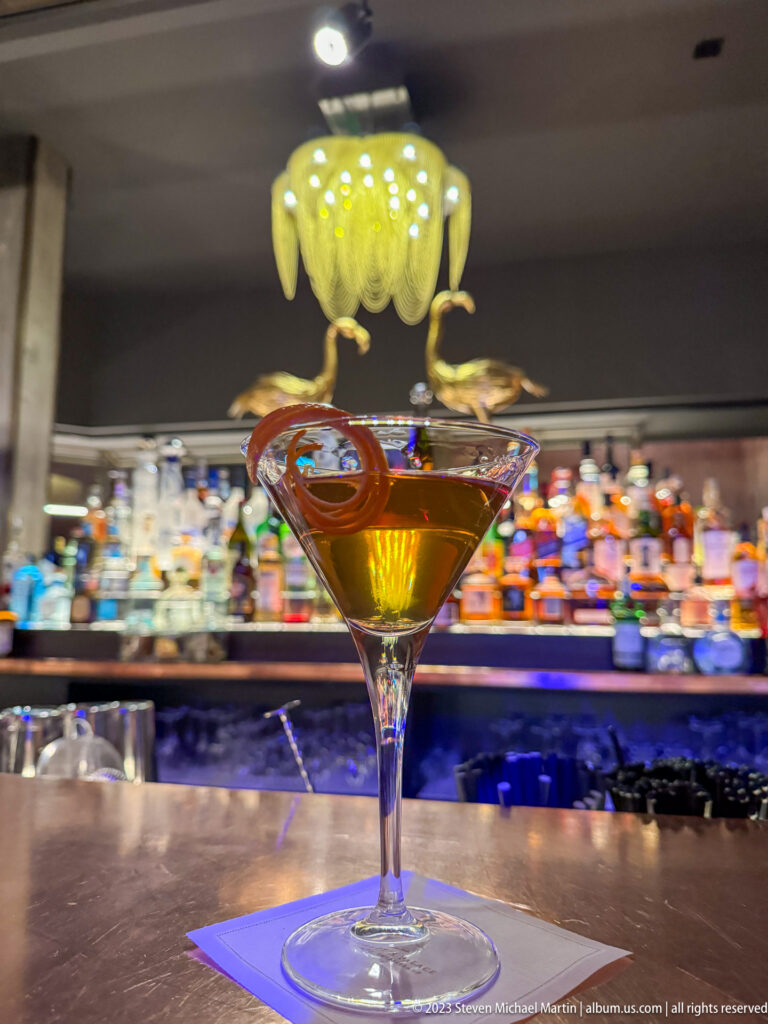 Amber cocktail in a wide stemmed glass with curled citrus peel, set before a blurred wall of colorful liquor bottles.