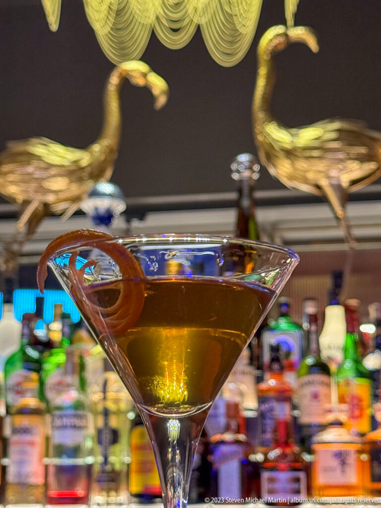 Close-up of an amber cocktail with curled citrus peel, colorful bottles behind, and two gold bird sculptures above the bar.