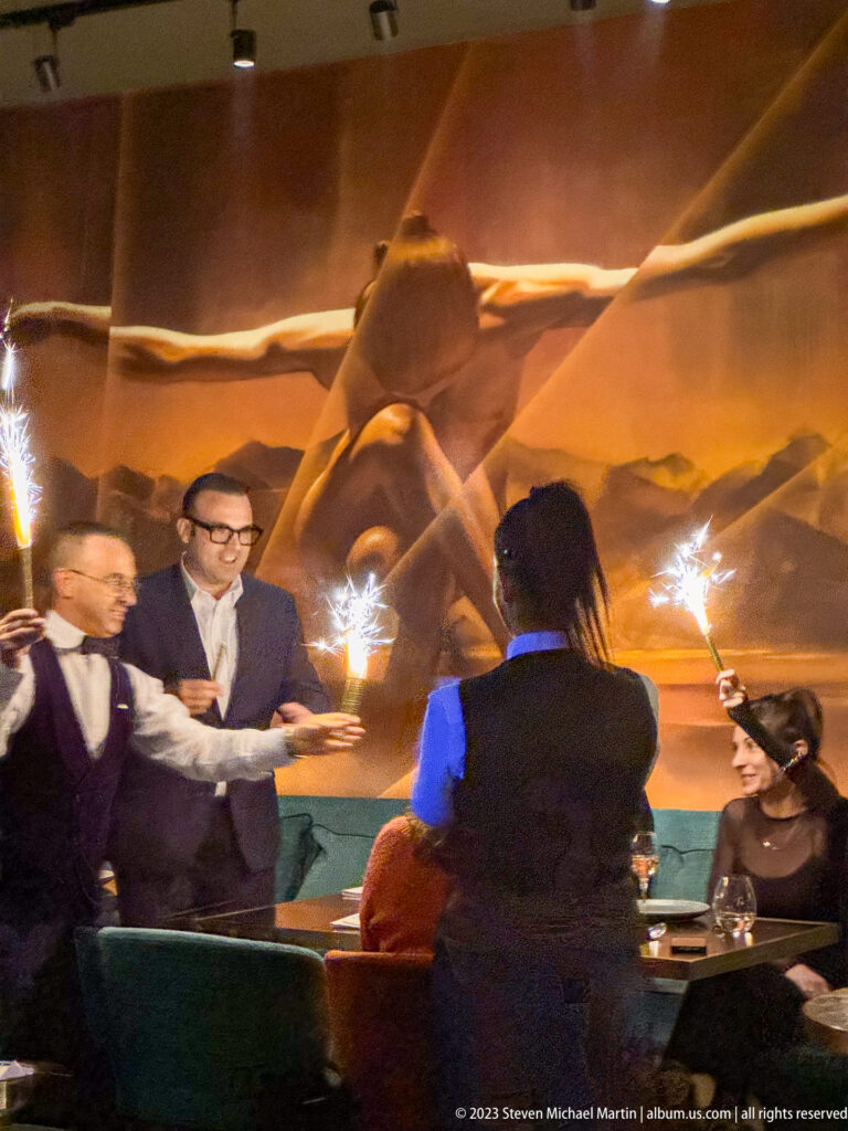 Restaurant staff hold lit sparklers around seated guests in a dim dining room with green banquettes and a mountain mural.