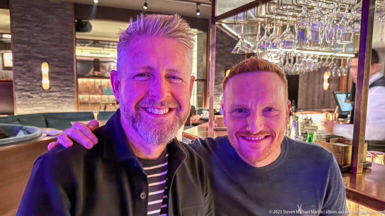 Two smiling men pose together beside a bar, with hanging glasses and softly lit restaurant furnishings behind them.