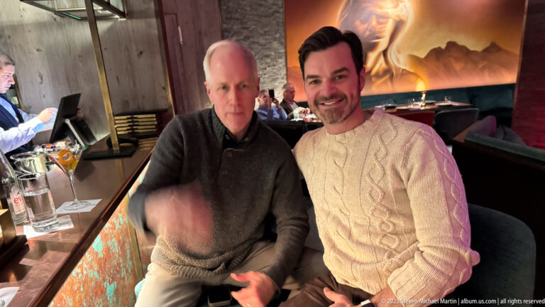 Two men seated together in a restaurant, one in dark gray and one in a cream cable-knit sweater, with another person partly visible.