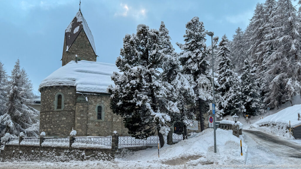Small stone church with a steep tower stands beside a snowy road among heavily snow-covered conifer trees.
